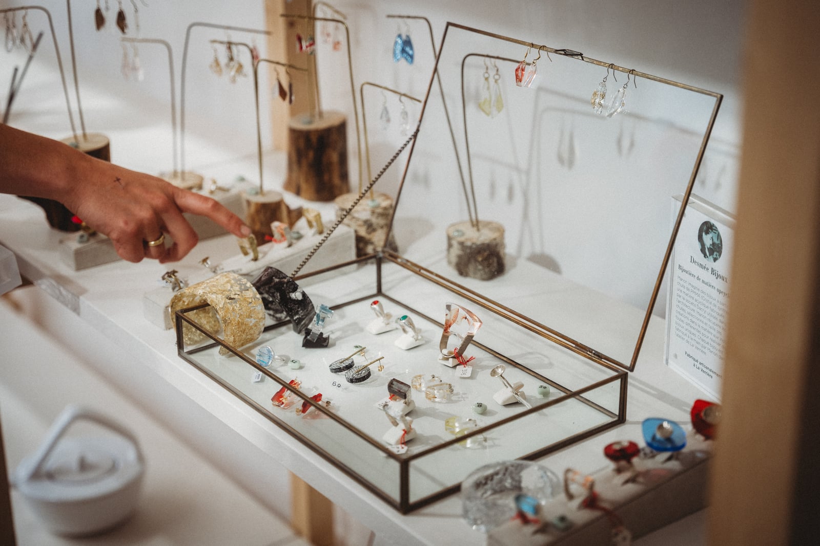 Coffret vitré présentant des bagues et boucles d'oreilles colorées sur un stand, une main désignant l'un des bijoux