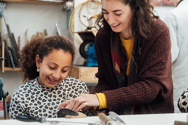 Une adulte souriante guide une enfant à l'établi, des outils à main posés sur le plan de travail
